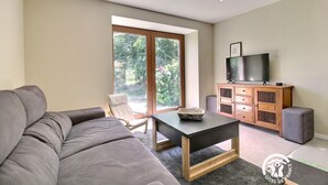 Ein modernes Wohnzimmer mit einem grauen Sofa, einem Holztisch, einem Fernseher auf einer Holzkommode und einer Glastür, die in einen Garten führt.