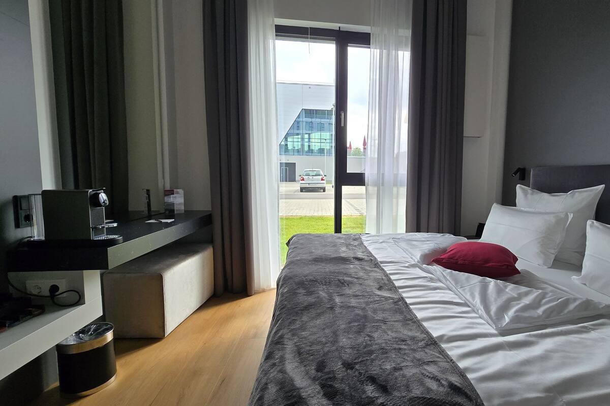 Een hotelkamer met een bed, een bureau met een computer, een prullenbak en uitzicht op een gebouw en een auto door het raam.