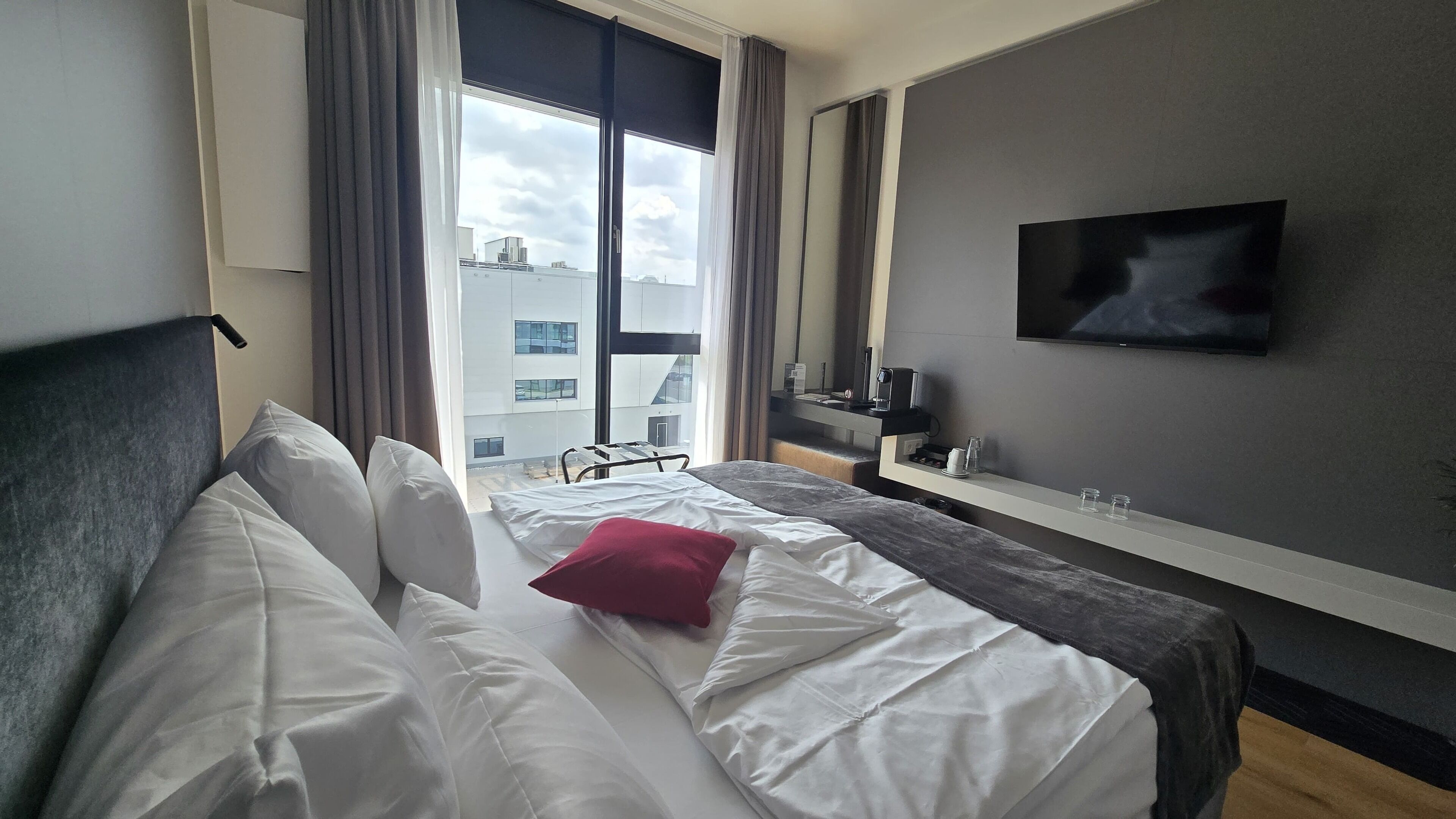 A hotel room with a bed, a TV, a desk, and a window with a view of buildings.