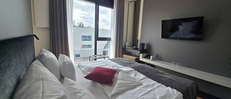 A hotel room with a bed, a TV, a desk, and a window with a view of buildings.