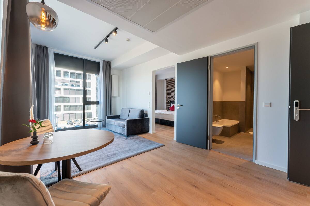 Een moderne hotelkamer met een houten vloer, een ronde eettafel, een bank en een groot raam met gordijnen.