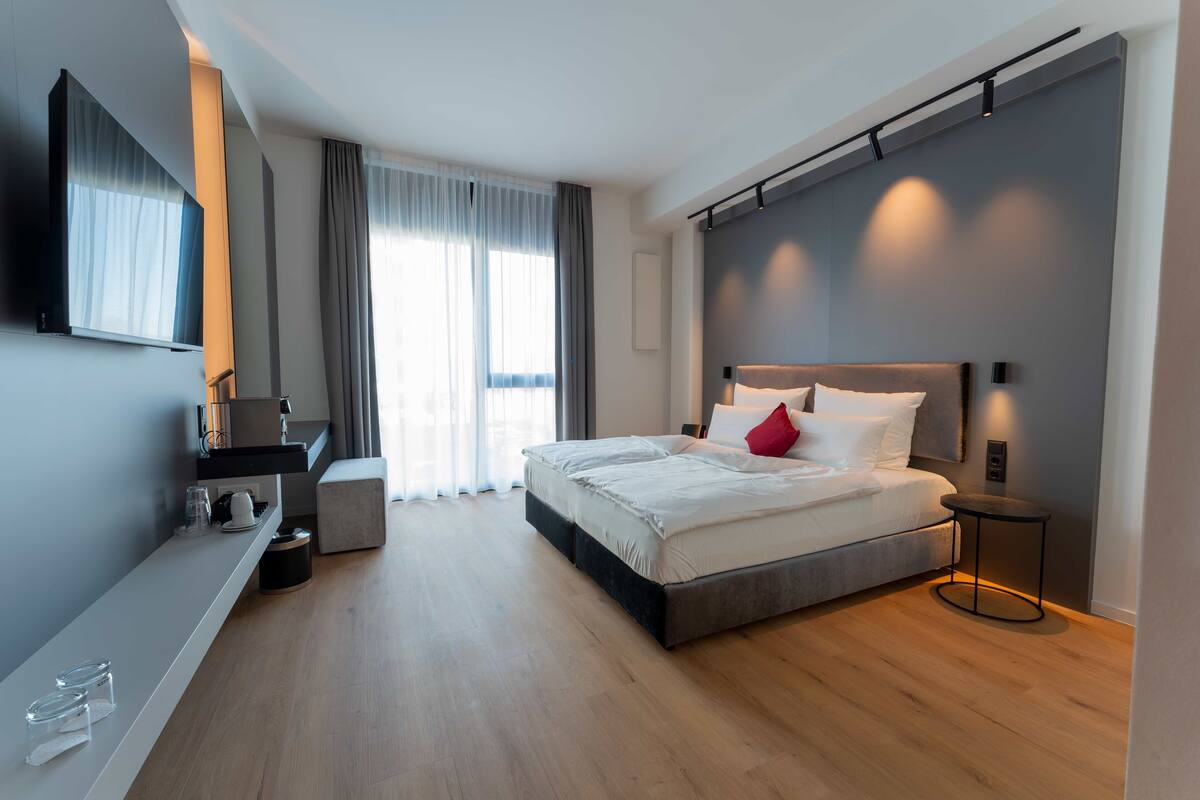 Een moderne hotelkamer met een groot bed, een nachtkastje, een flatscreen televisie en een groot raam met gordijnen.