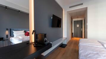 A modern hotel room with a large bed, a desk with a coffee maker, a flat-screen TV mounted on the wall, and a wooden floor.