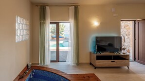 A room with a wooden bathtub, a television on a wooden stand, and a view of a pool through a glass door. - Istros Villa III, Infinite Blue, Pool & Whirlpool (G37Q+HX Pithari, Greece)