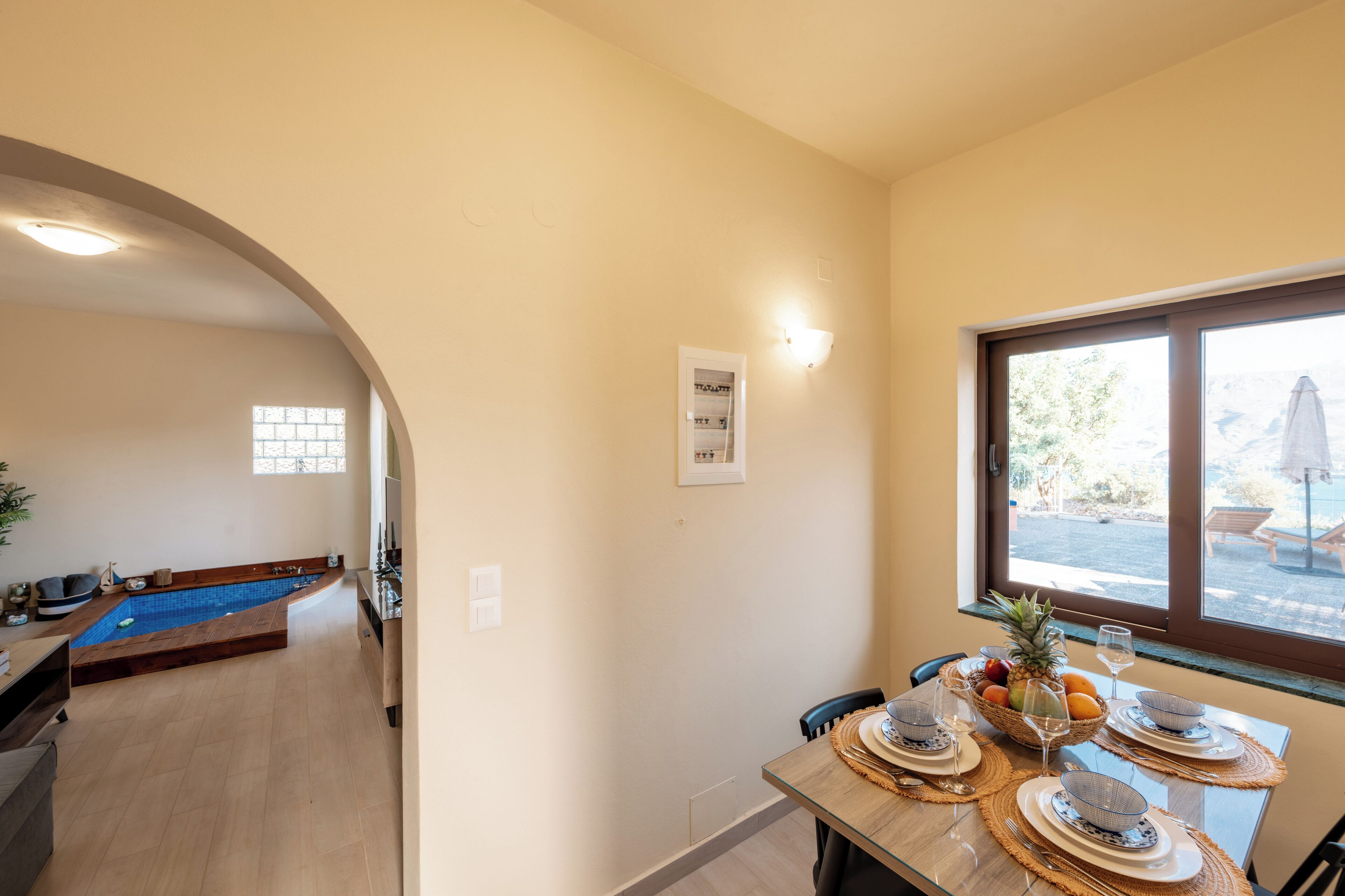 A dining area with a pool in the background, a window with a view, and a table set for two with plates, glasses, and a fruit basket.