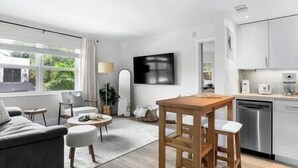 A modern living room with a sofa, a round coffee table, and a dining table. There is a kitchen area with cabinets and a dishwasher. - Cozy corner condo, just renovated,  pool views (Fort Lauderdale)
