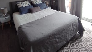 Une chambre moderne avec un grand lit, une table de chevet, une commode, une étagère murale et une fenêtre avec des rideaux.