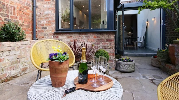 Een terras met een rieten stoel, een tafel met een fles en glazen, en bloempotten.