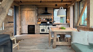 Interior rústico de una cabaña con piso de madera, una zona de comedor con mesa y sillas, una cocina con armarios de madera y estufa, y ventanas grandes.