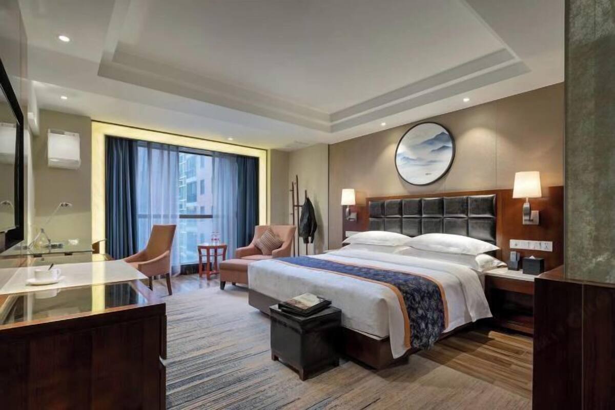 A hotel room with a bed, bedside tables, a desk, a chair, a small table, and a large window with curtains.