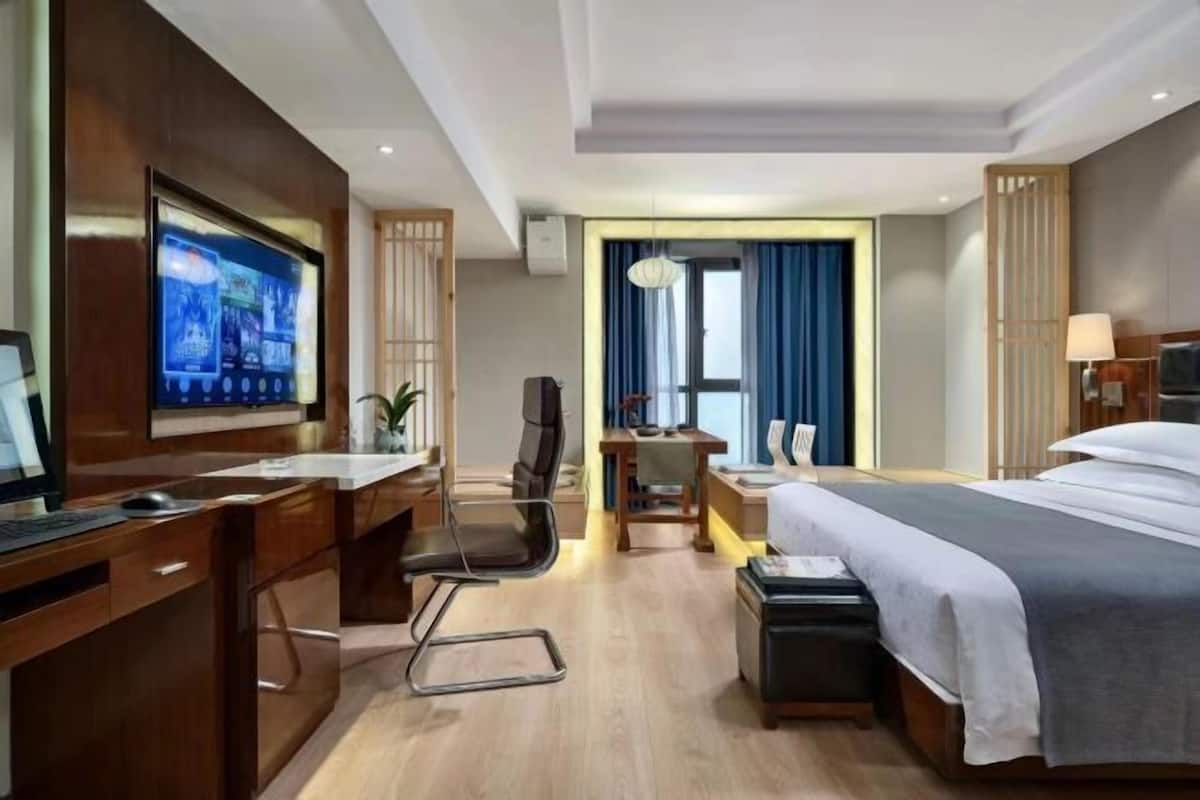 A hotel room with a bed, desk, chair, TV, and a view of the outside.