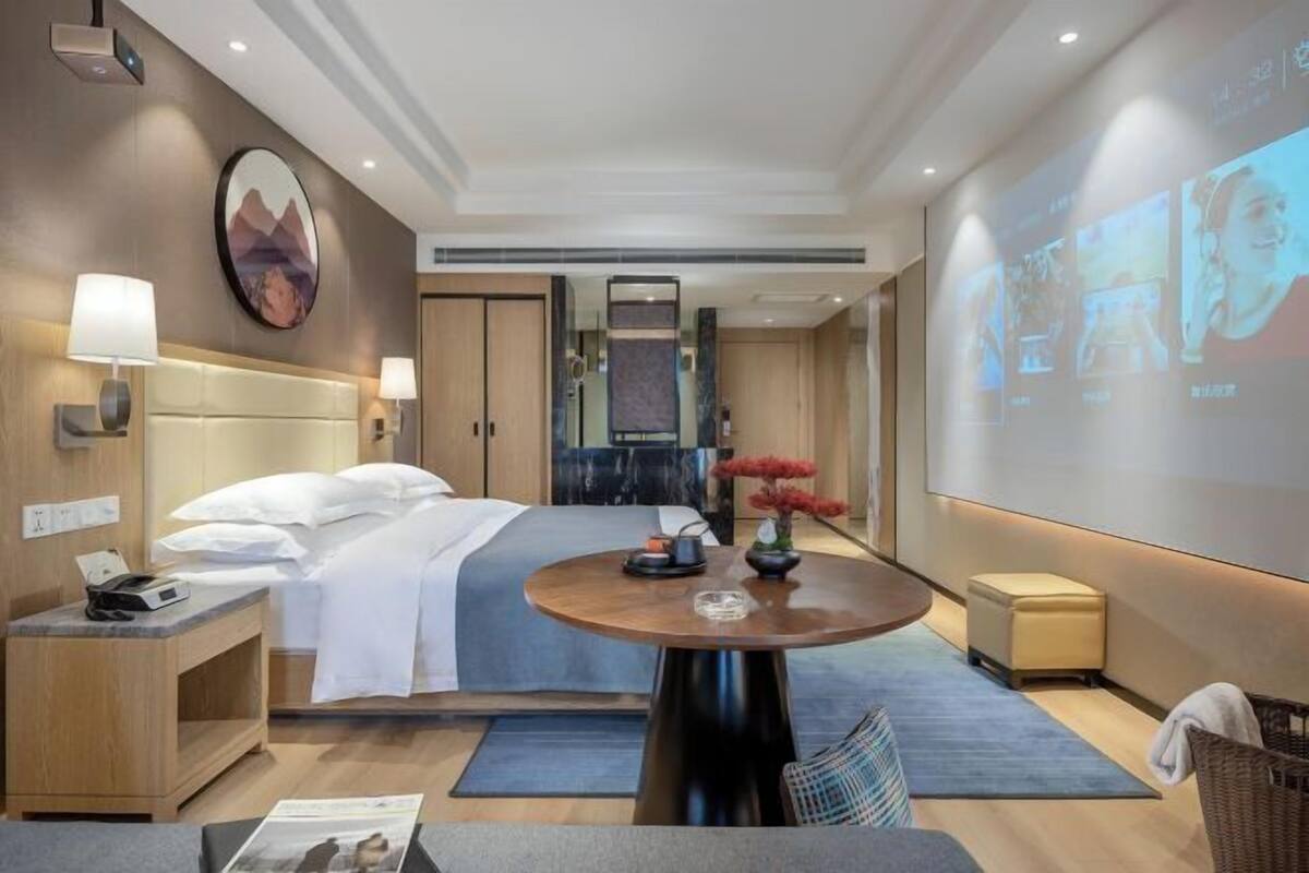 A hotel room with a bed, bedside tables, a round table, a wall-mounted projector, and a large screen.
