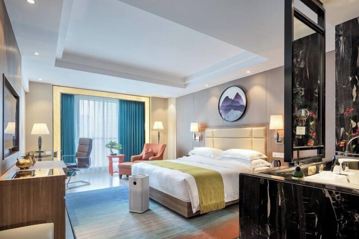 A hotel room with a large bed, a desk, a chair, a small table, a plant, and a bathroom with a mirror and sink.