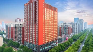 A high-rise building with a red facade, surrounded by a tree-lined street and other buildings in the background.