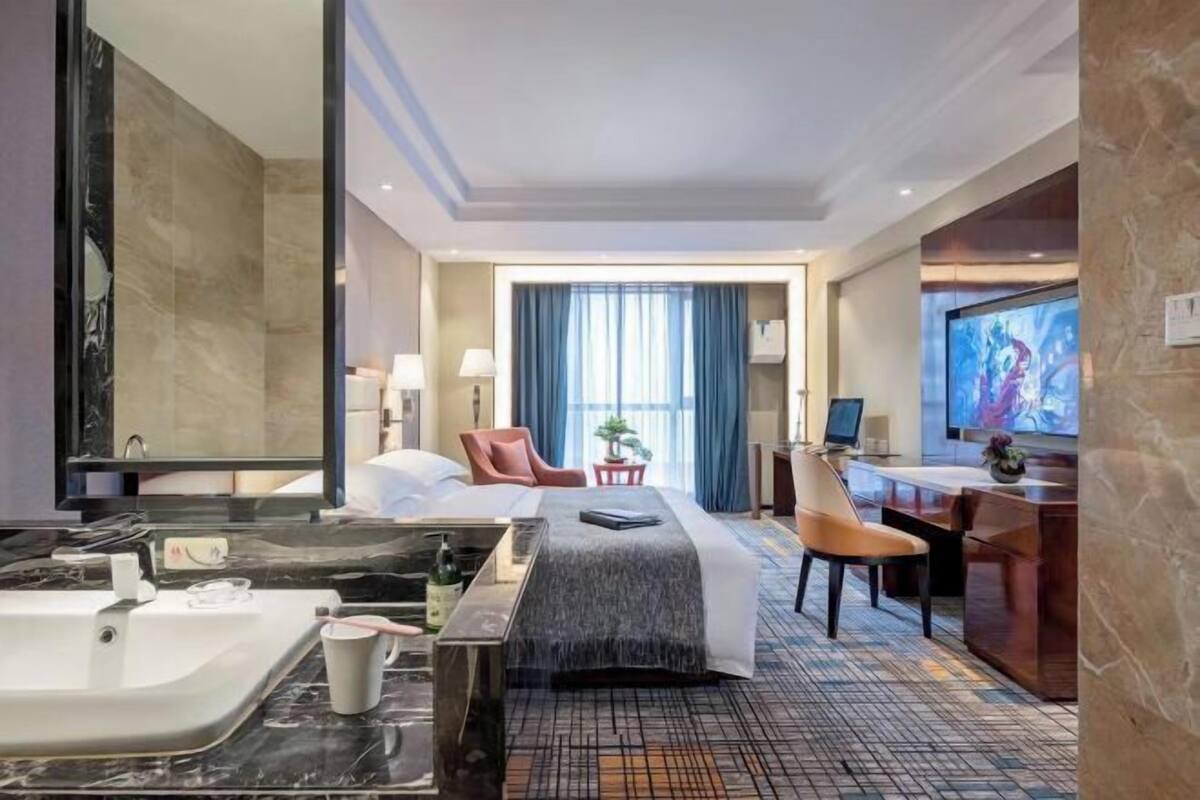 A hotel room with a large bed, a desk, a chair, a TV, a bathroom with a bathtub, and a large window with curtains.
