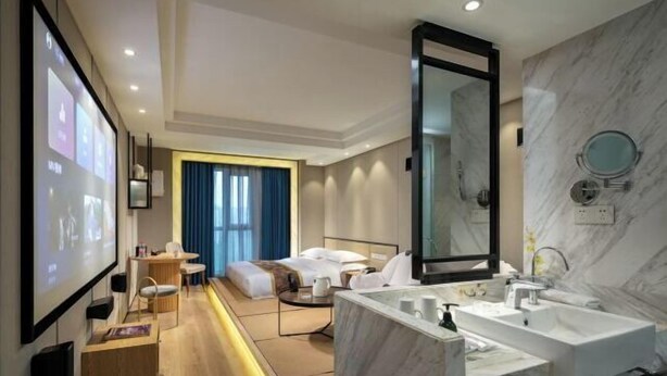 A modern hotel room with a large bed, a sitting area with a table and chairs, a large bathroom with a marble sink and mirror, and a projection screen on the wall. - Ming Yi Hotel (Zhuzhou)