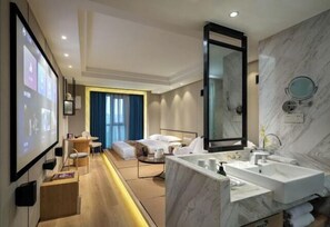 Ein modernes Hotelzimmer mit einem großen Bett, einer Sitzecke mit Tisch und Stühlen, einem geräumigen Badezimmer mit Marmorwaschbecken und Spiegel sowie einer projizierten Leinwand an der Wand.