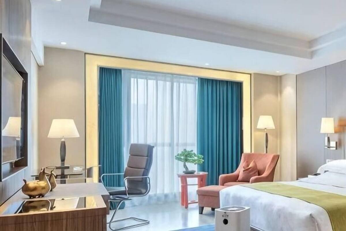 A hotel room with a bed, two chairs, a desk, a lamp, a small table with a plant, and a window with curtains.