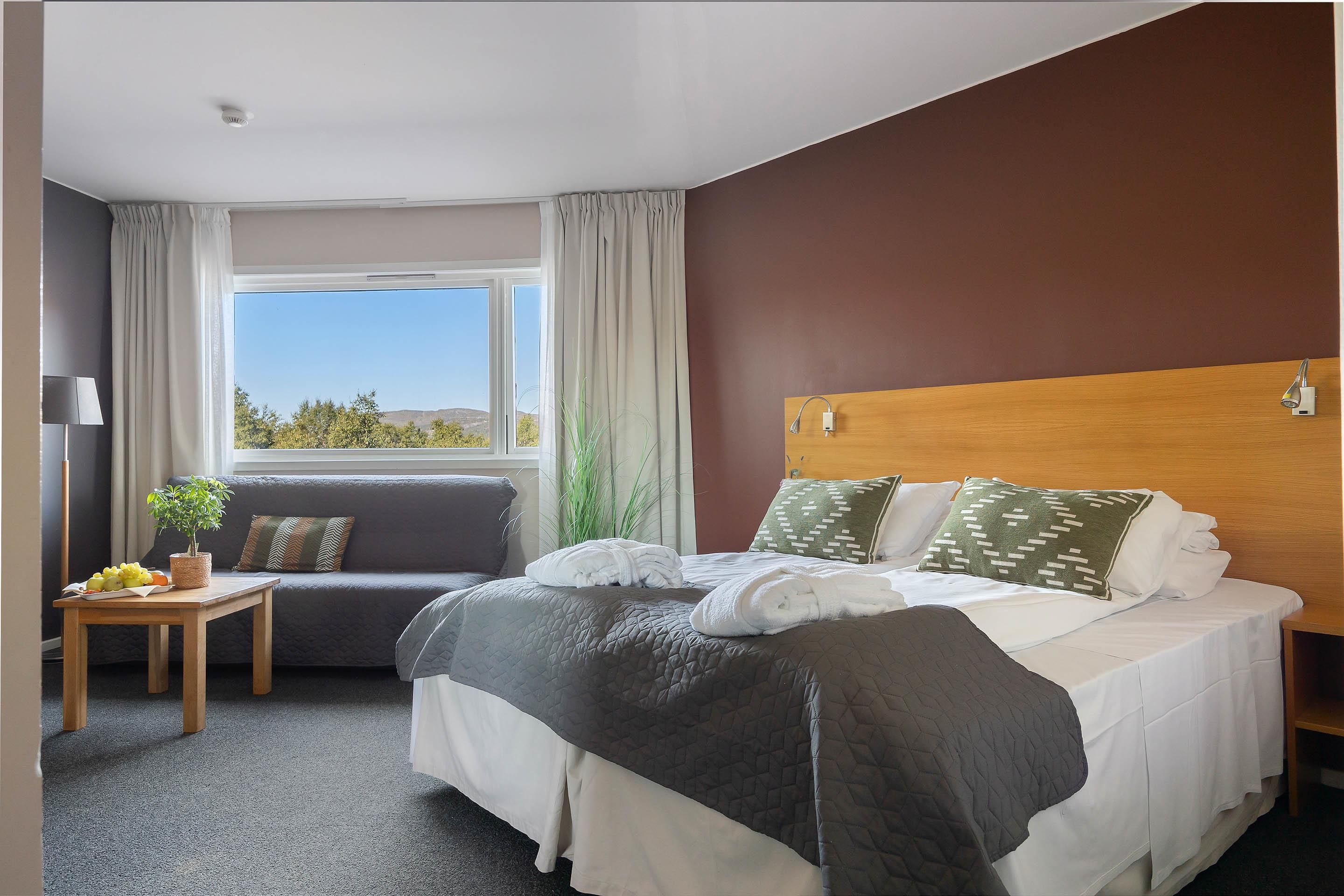 Una habitación de hotel con cama, sofá, una mesita con una planta y ventana con vistas al exterior.