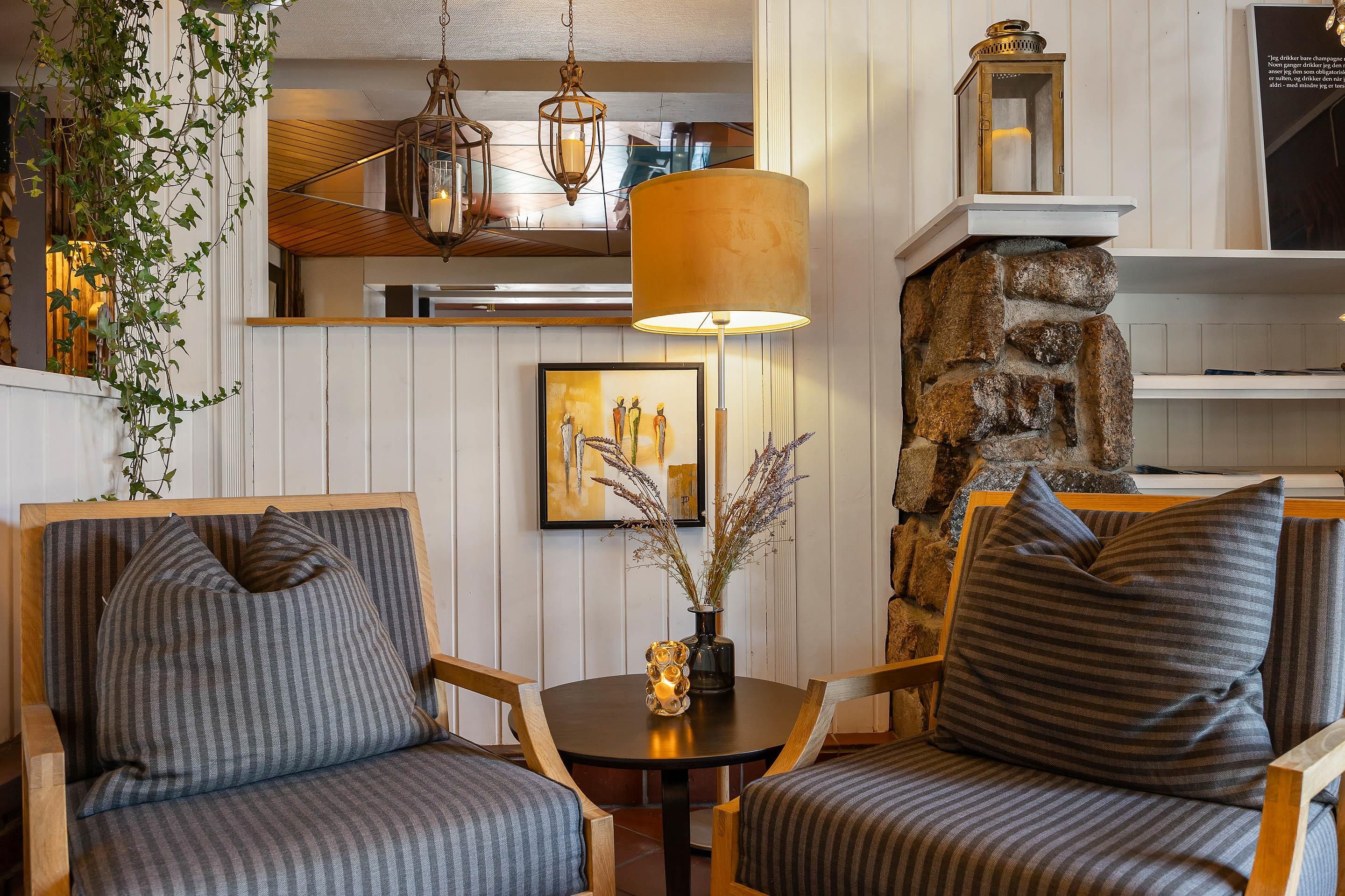 Un coin salon confortable avec deux fauteuils, une table et une lampe. Un tableau encadré orne le mur, et un vase décoratif rempli de fleurs est posé à proximité.