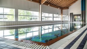 Une piscine intérieure avec des parois en verre transparent, des poutres en bois et un revêtement de sol carrelé.