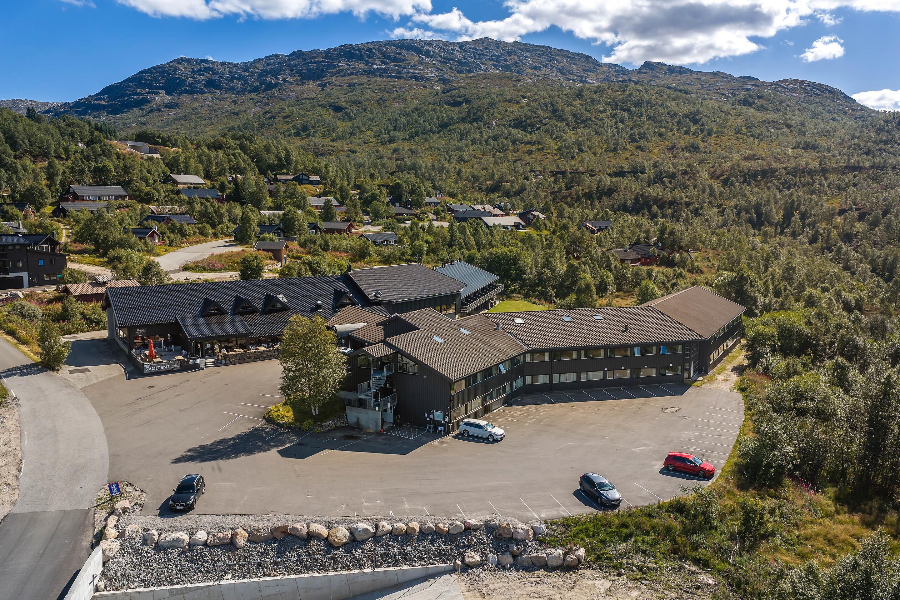 A modern building with multiple parking spaces and a surrounding landscape with trees and hills.