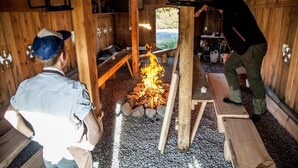 L’intérieur d’une cabane en bois, avec un foyer central, des bancs et une table.