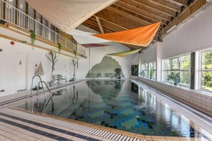 An indoor swimming pool with a tiled floor, a diving board, and a view of the outdoors through large windows.