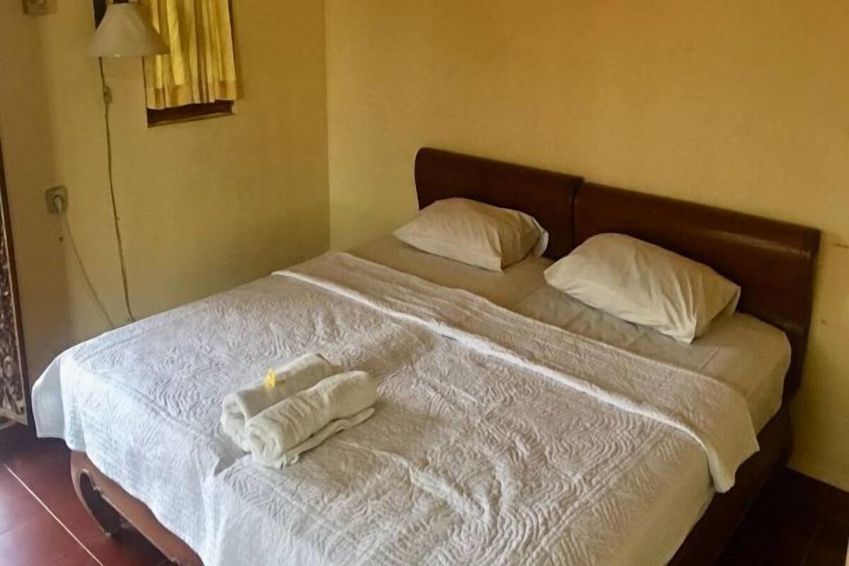A neatly made bed with white linens and two pillows, a bedside lamp, and a window with yellow curtains.