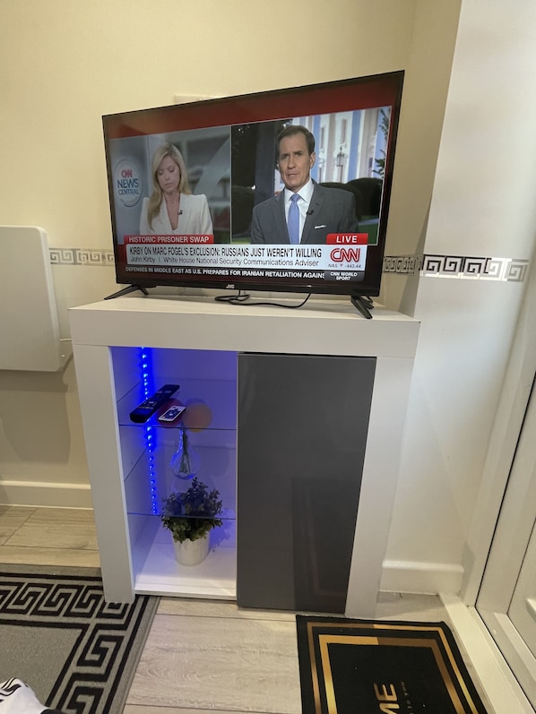 A television on a white stand displaying a news program. Below the TV, there is a glass shelf with a remote control, a vase with flowers, and a decorative item.