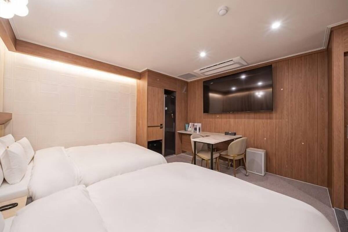 Ein Hotelzimmer mit zwei Betten, einem Schreibtisch, einem Sessel, einem Fernseher und einer Wandpaneele aus Holz.