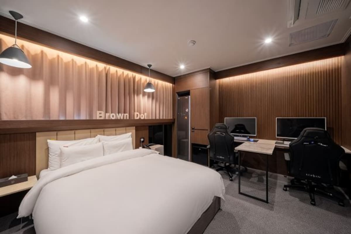 A hotel room with a large bed, a desk with two monitors, and a gaming chair.