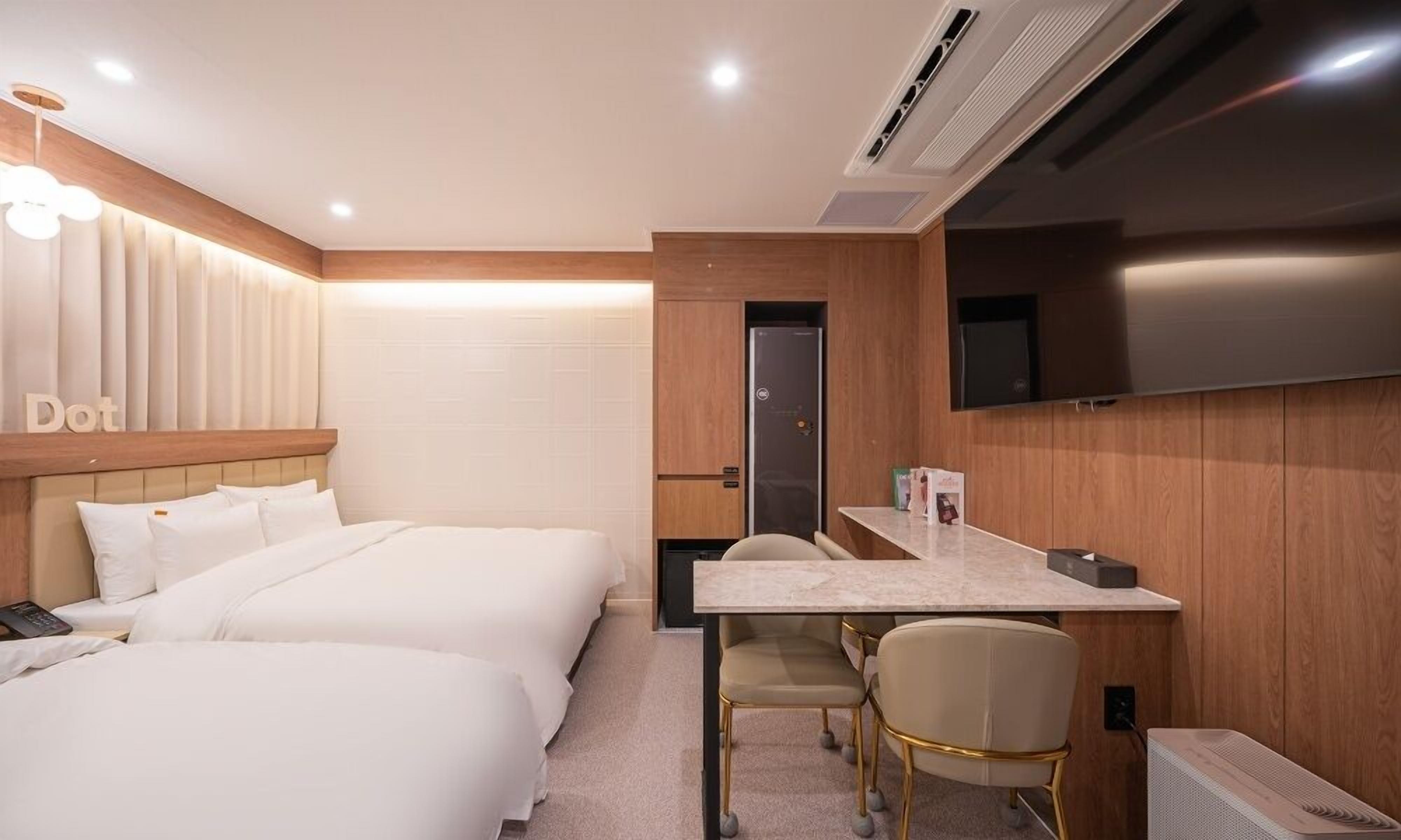 A hotel room with two beds, a desk, a chair, a TV, and an air conditioning unit.