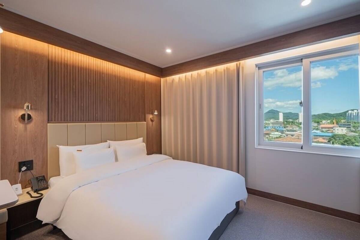 Ein Hotelzimmer mit einem großen Bett, einem Nachttisch, einer Wandlampe und einem Fenster mit Blick auf die Stadt.