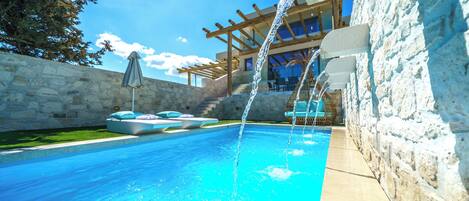 A swimming pool with a stone wall and a pergola with a water feature.