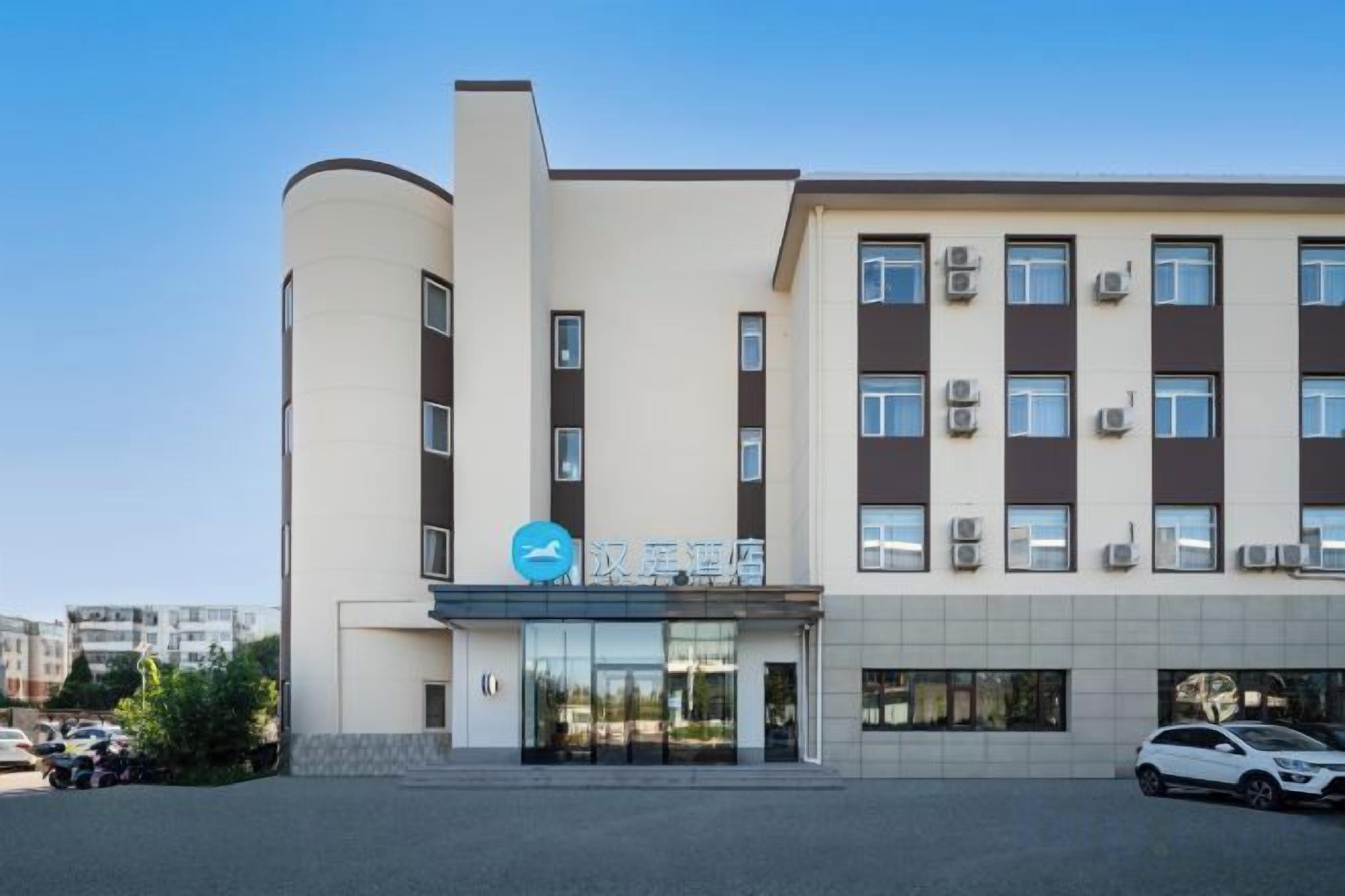 Un hotel de varios pisos con fachada moderna, una entrada destacada con un logotipo azul y un área de estacionamiento al frente.