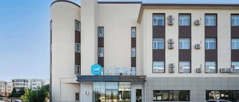 Un hotel de varios pisos con fachada moderna, una entrada destacada con un logotipo azul y un área de estacionamiento al frente.