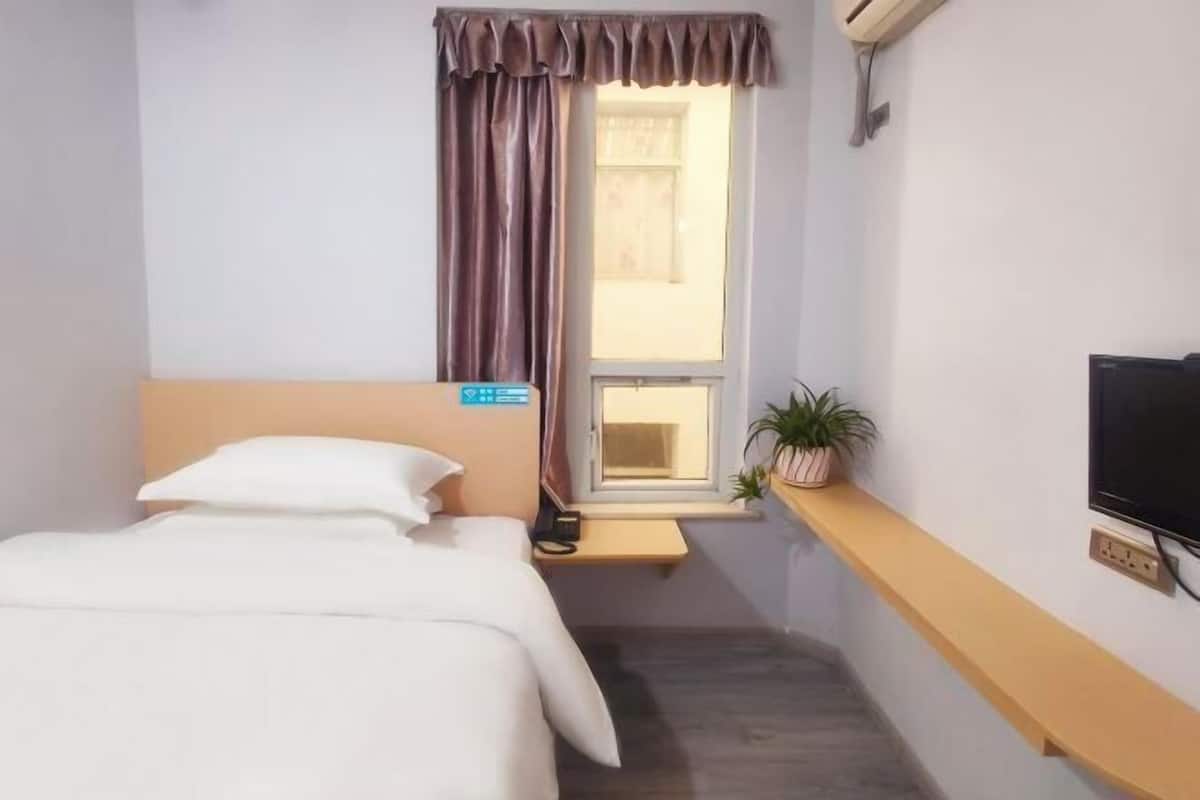 Une chambre d’hôtel avec un lit, une fenêtre avec des rideaux, une petite étagère avec une plante et un téléviseur fixé au mur.