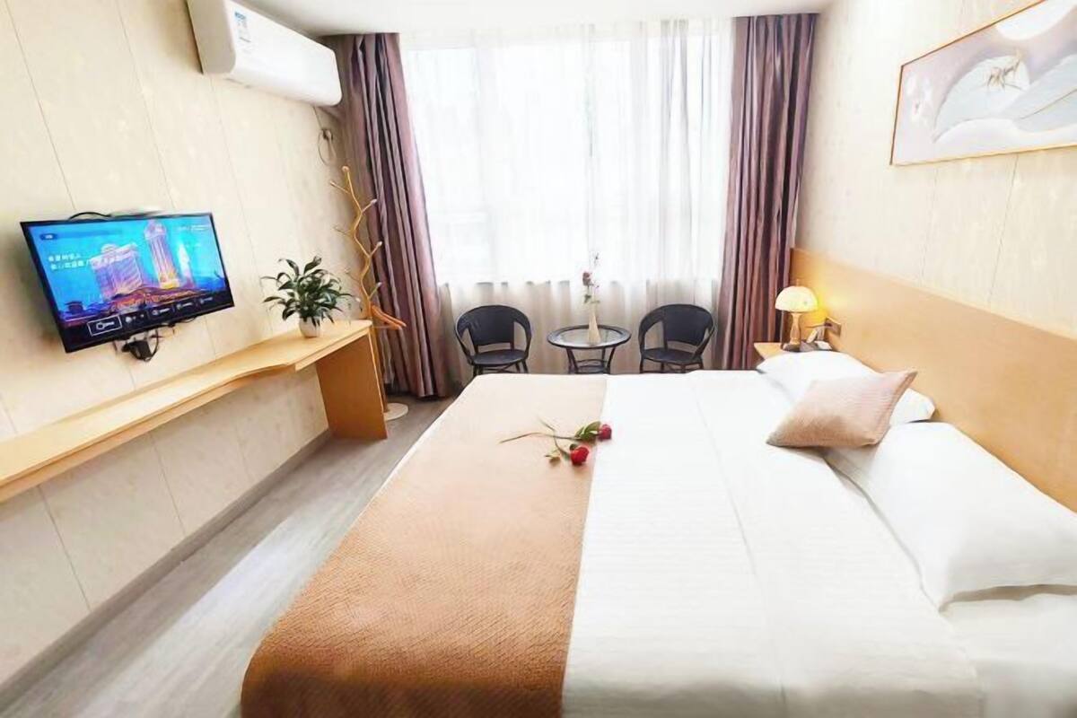Une chambre d’hôtel avec un lit, un téléviseur fixé au mur, un bureau agrémenté d’une plante, deux chaises, une petite table et une lampe.