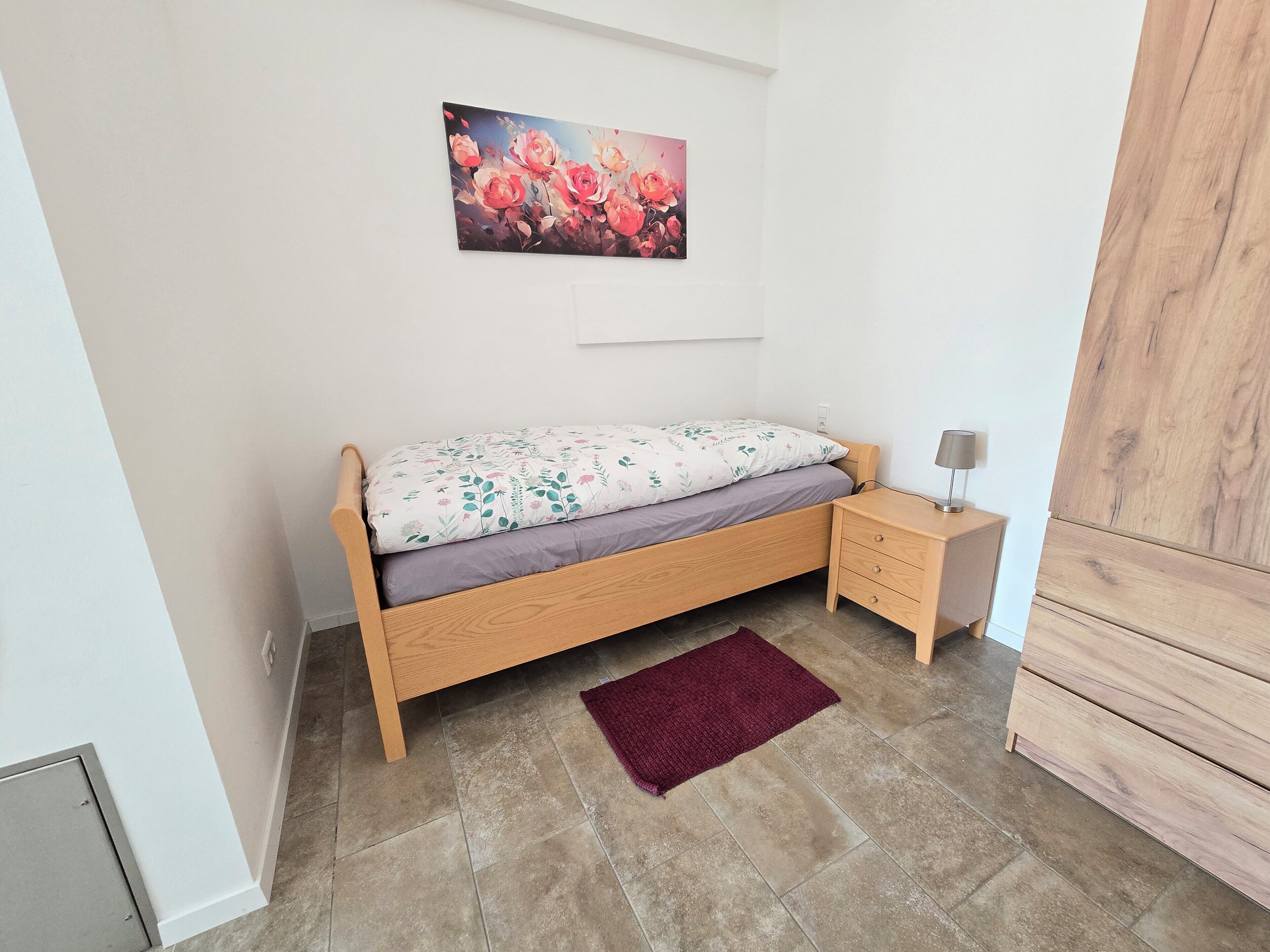 Ein Einzelbett mit floralem Überwurf, ein Nachttisch mit Lampe, ein roter Teppich und ein Blumenbild an der Wand.