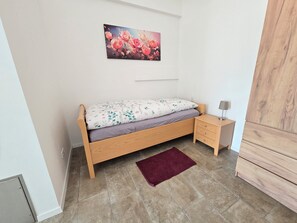 Un lit simple avec une couvre-lit à motifs floraux, une table de chevet avec une lampe, un tapis rouge et un tableau représentant des fleurs accroché au mur.
