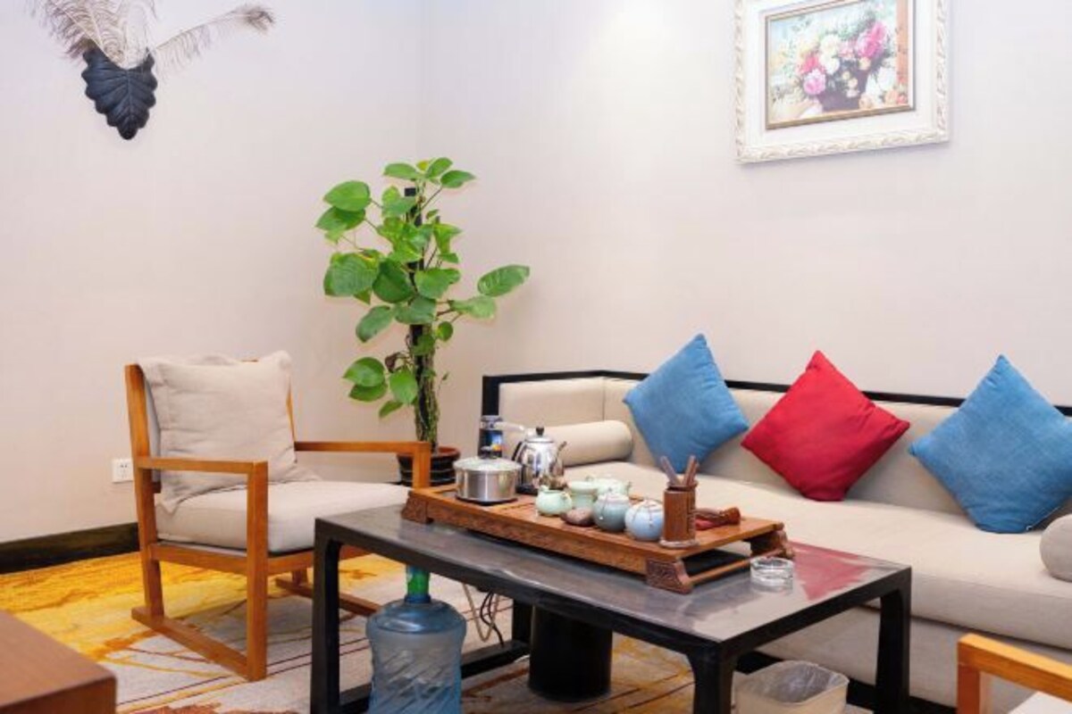 A living room with a sofa, a wooden chair, a coffee table with teapots, a potted plant, and a wall-mounted decoration.