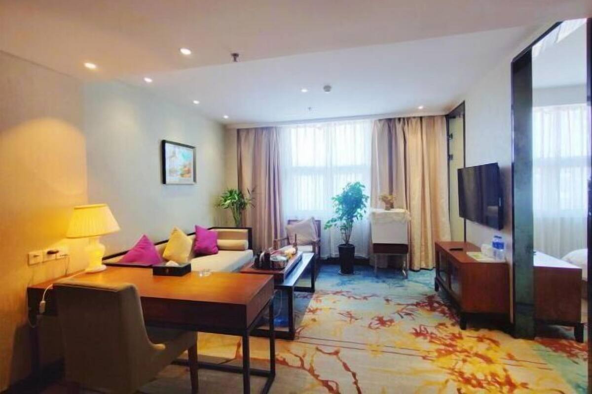 A hotel room with a bed, a desk, a chair, a TV, a lamp, a plant, and a window with curtains.