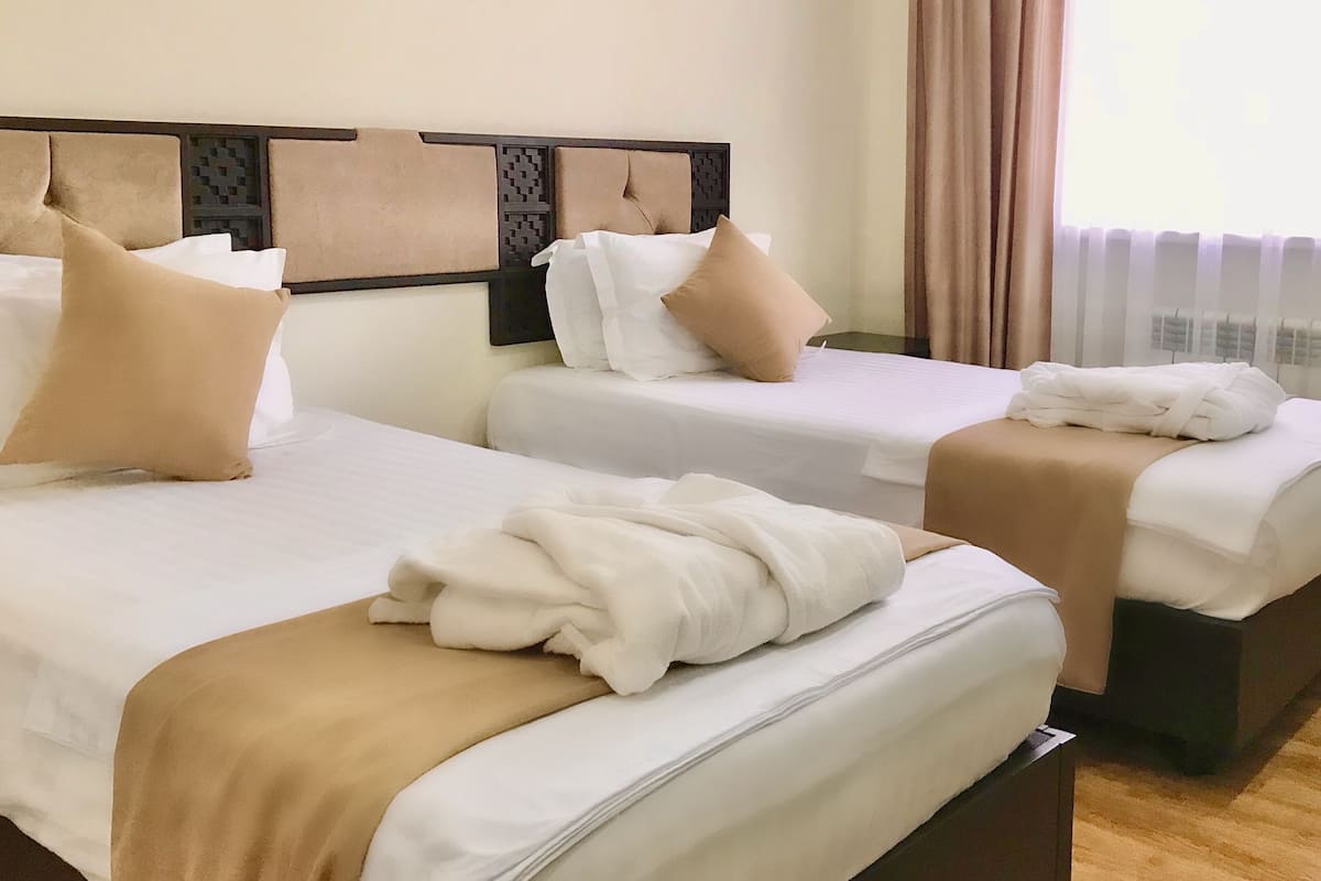 Habitación de hotel con dos camas, ropa de cama color beige y blanco, cabecero con diseño geométrico y una ventana con cortinas.