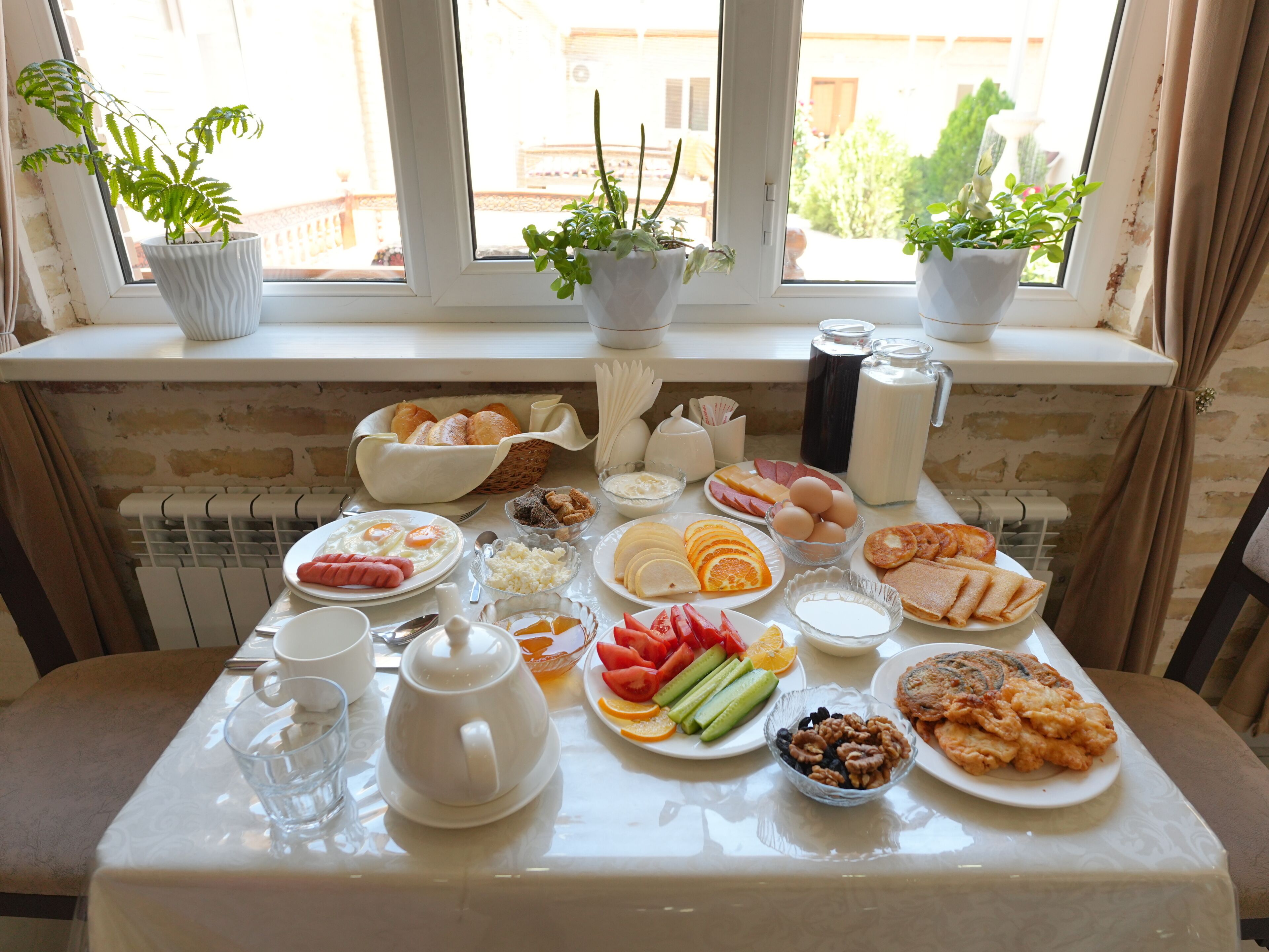 A breakfast table with various food items, a teapot, and potted plants on a windowsill.