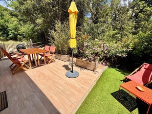 Une terrasse en bois avec une table et des chaises, un parasol jaune et un petit jardin avec de lâherbe et des plantes.