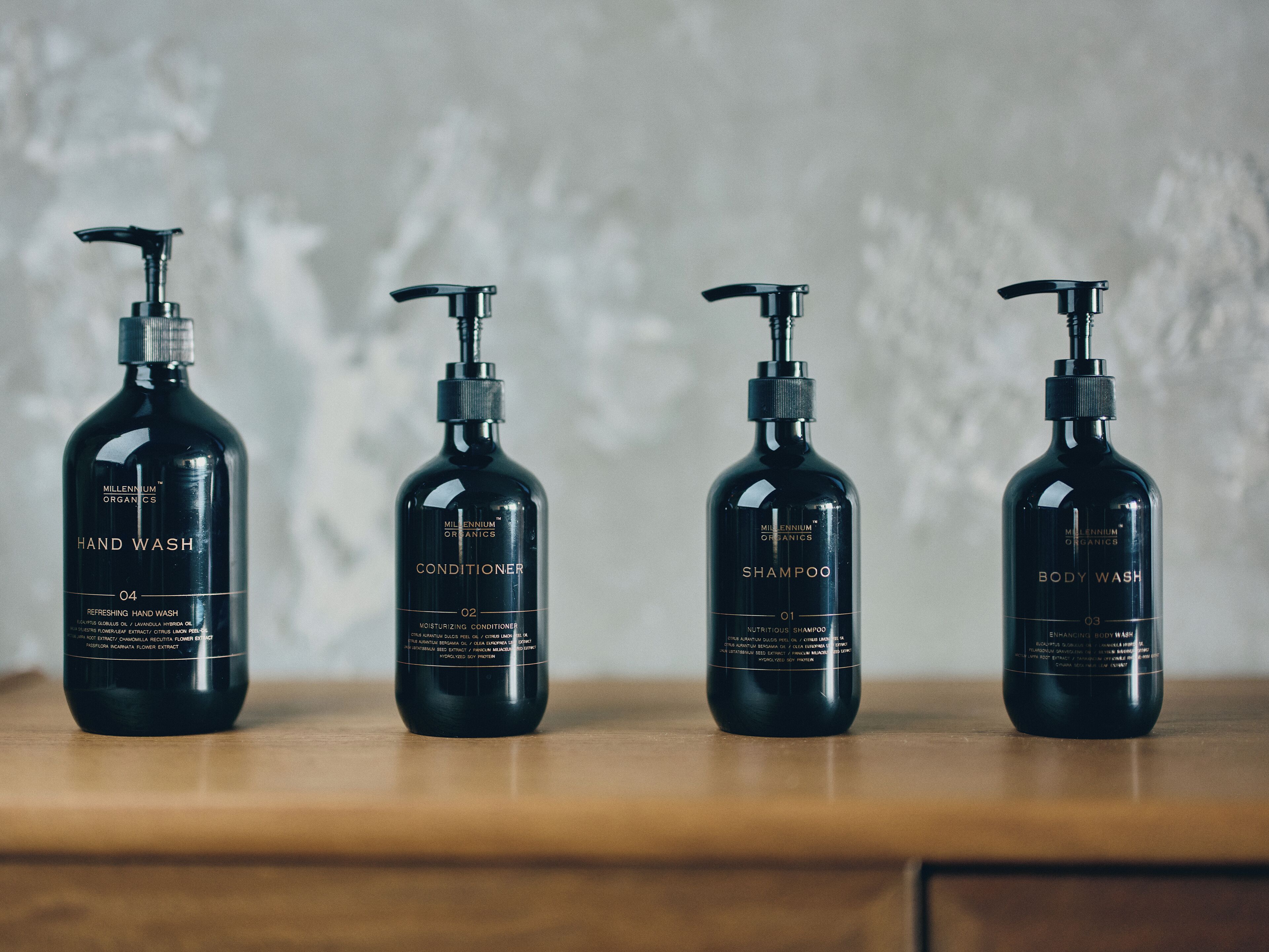Quatre bouteilles noires de la marque Millennial Organics contenant du savon pour les mains, de l’après-shampoing, du shampoing et du gel douche, disposées sur une surface en bois.