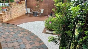 A patio area with a brick pathway, a wooden deck, and a seating arrangement with chairs and a table. - Gästehaus am Ruppiner See (Neuruppin)