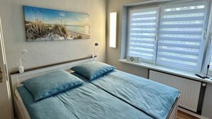 A bedroom with a bed, a painting, a window with blinds, and a bedside table. - Gästehaus am Ruppiner See (Neuruppin)