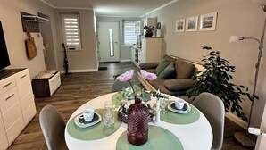 A dining area with a round table, chairs, and a vase with flowers. There is a sofa and a plant in the background. - Gästehaus am Ruppiner See (Neuruppin)